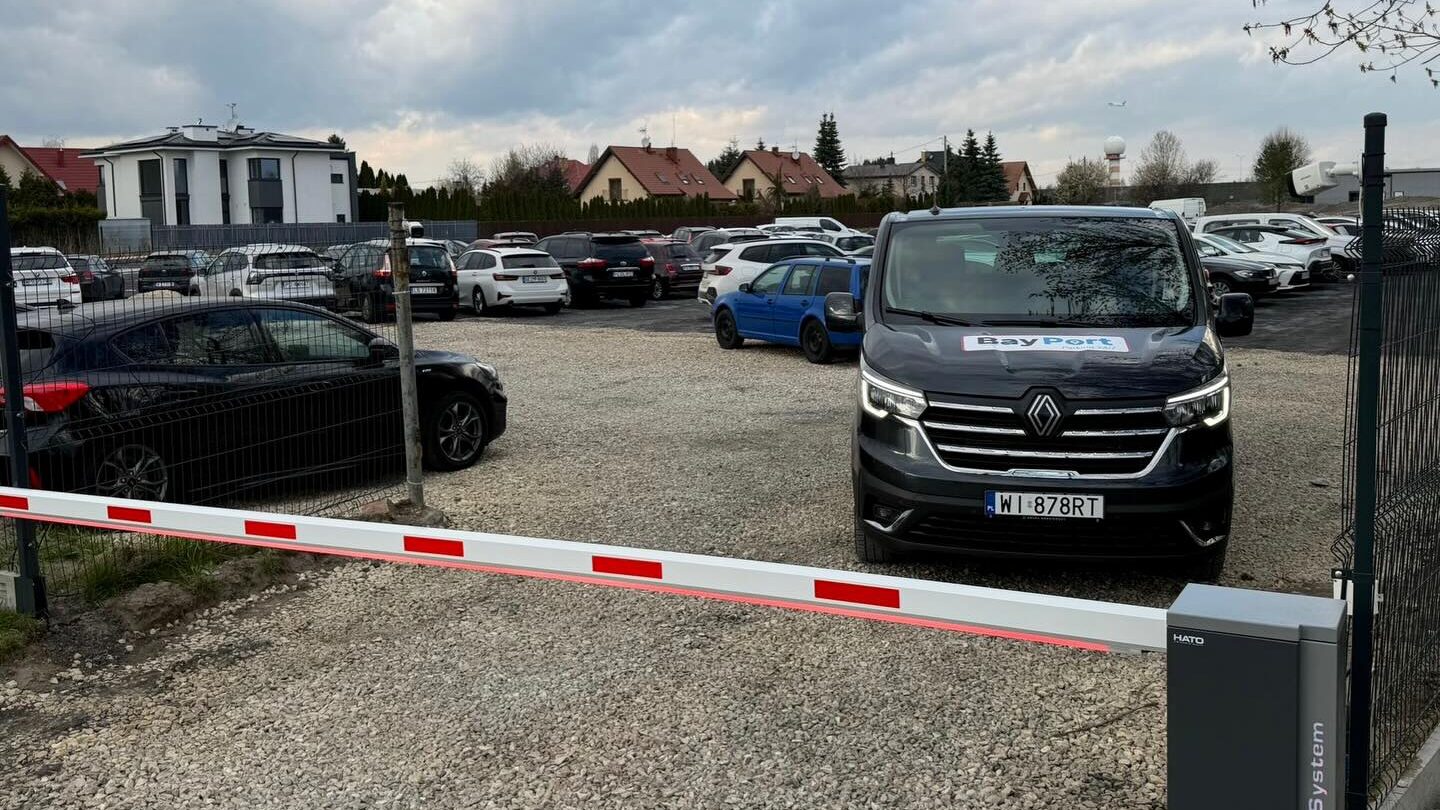 Czarny bus BayPort przy szlabanie wjazdowym na parking w Warszawie, w otoczeniu zaparkowanych samochodów osobowych.